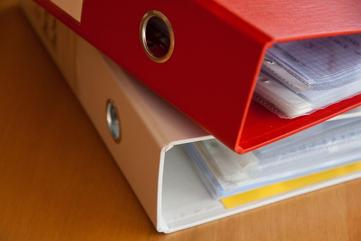Close-up of a red and a white office binder stacked on a wooden desk, with plastic sleeves of papers inside