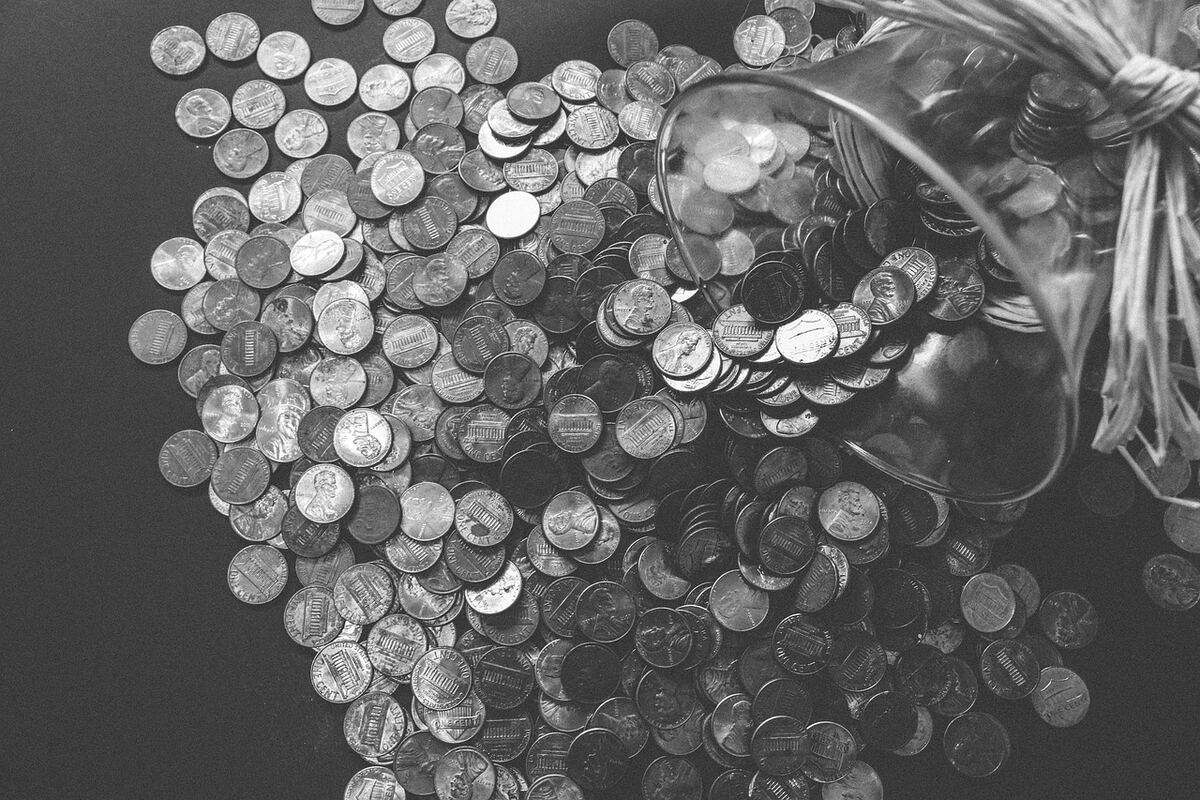 Black-and-white overhead photo of a glass jar tipped over, with coins spilling across a dark surface